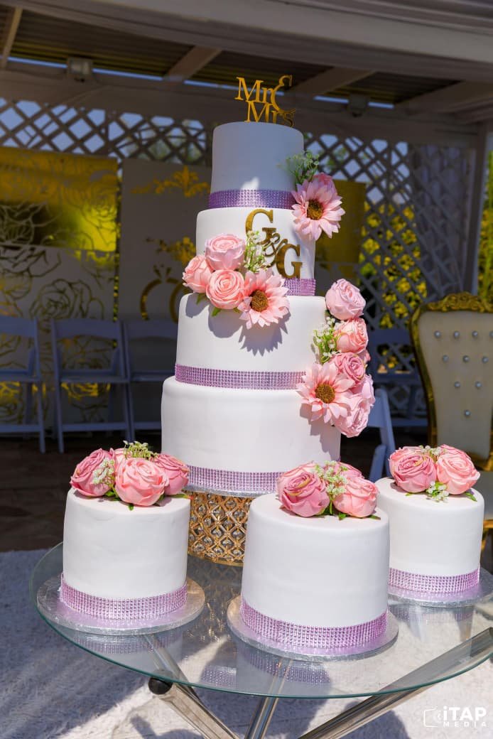 Four-tier white wedding cake with pink roses and Mr & Mrs gold topper, surrounded by matching mini cakes